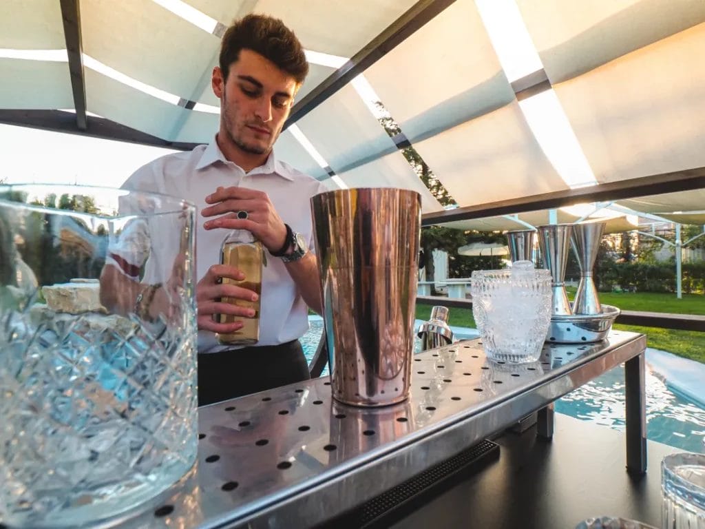 A mobile bartender wearing a white button-up shirt holds a bottle of liquor at a drinks stand under a canopy at a wedding.