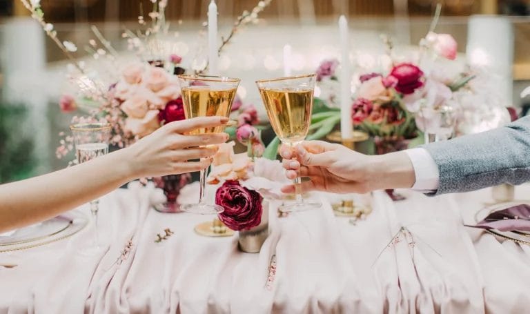 Close-up of a bride and groom having a toast with flutes of champagne at their reception table full of pink and burgundy flowers.