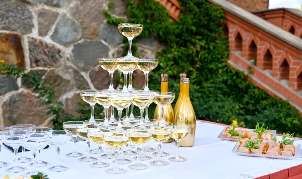 A champagne tower sits on a reception table next to some appetizers at an outdoor venue.