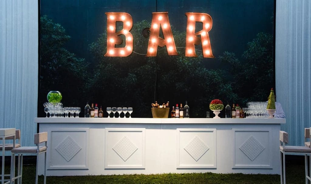Light up letters that spell out "BAR" hang above a white bar setup at an indoor venue for a wedding.