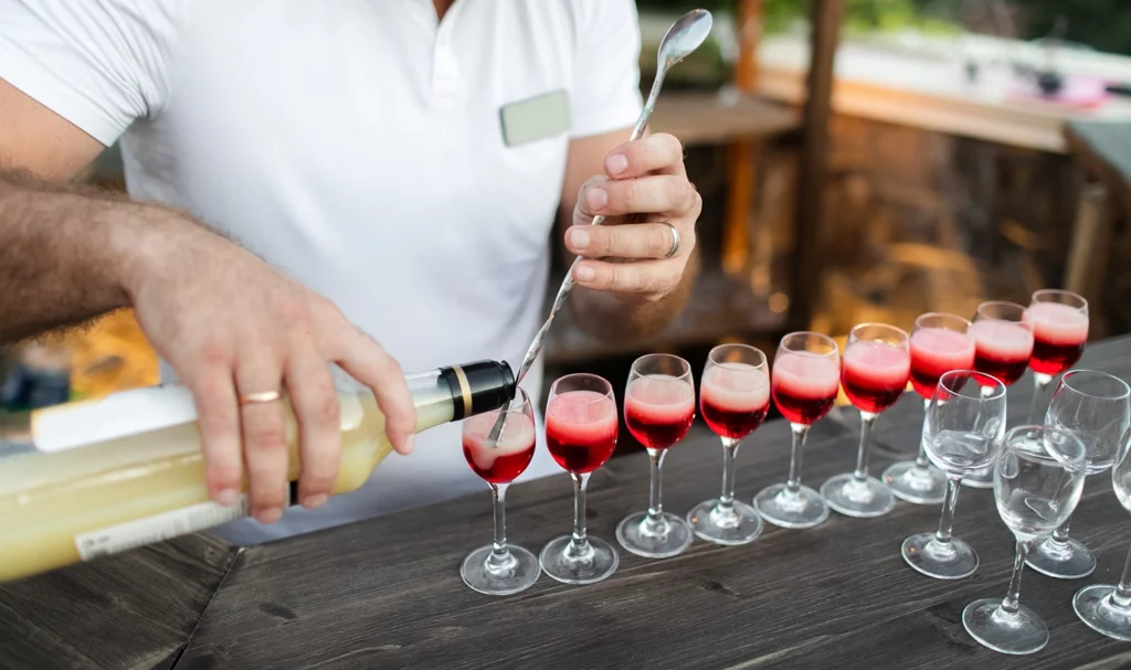 A bartender is using the handle of a long spoon to control his pour and layer alcoholic drinks in small cocktail glasses.