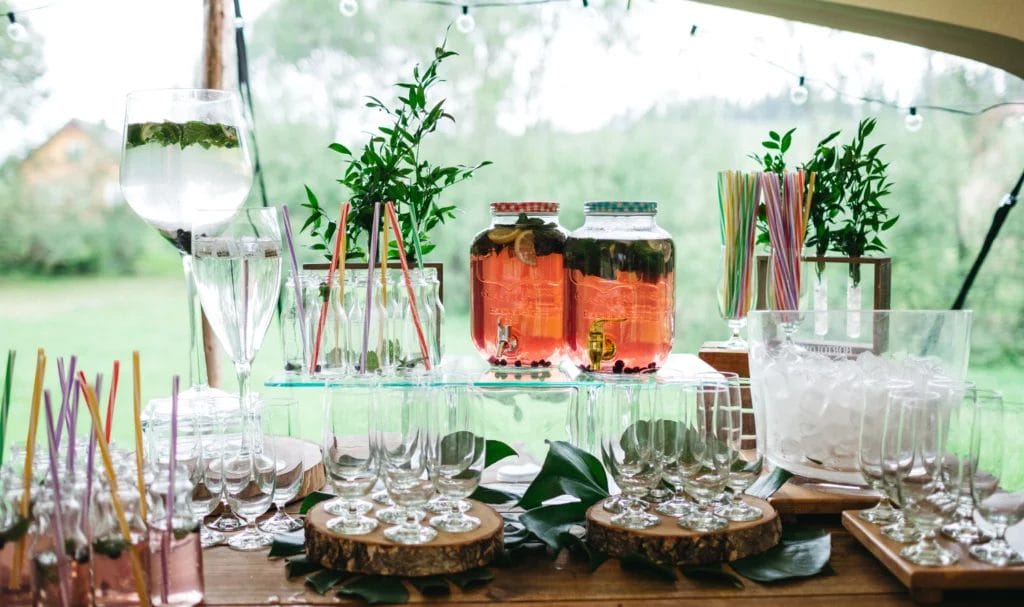 Front view of a drink bar at an outdoor wedding party with empty glasses, prepared drinks, full drink dispensers, and ice buckets neatly arranged on a wooden table.