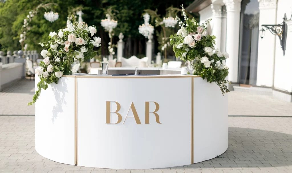 Bar counter with the large words "BAR" in all caps on the front, is decorated with white flowers at a wedding party.