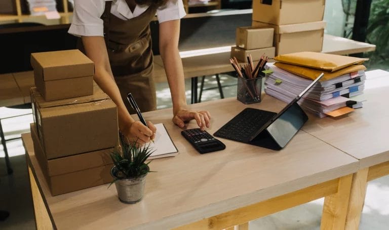 Close-up of a small business owner working from a home office. They are using a calculator while taking down notes on a desk next to a tablet, paperwork, and boxes of inventory.