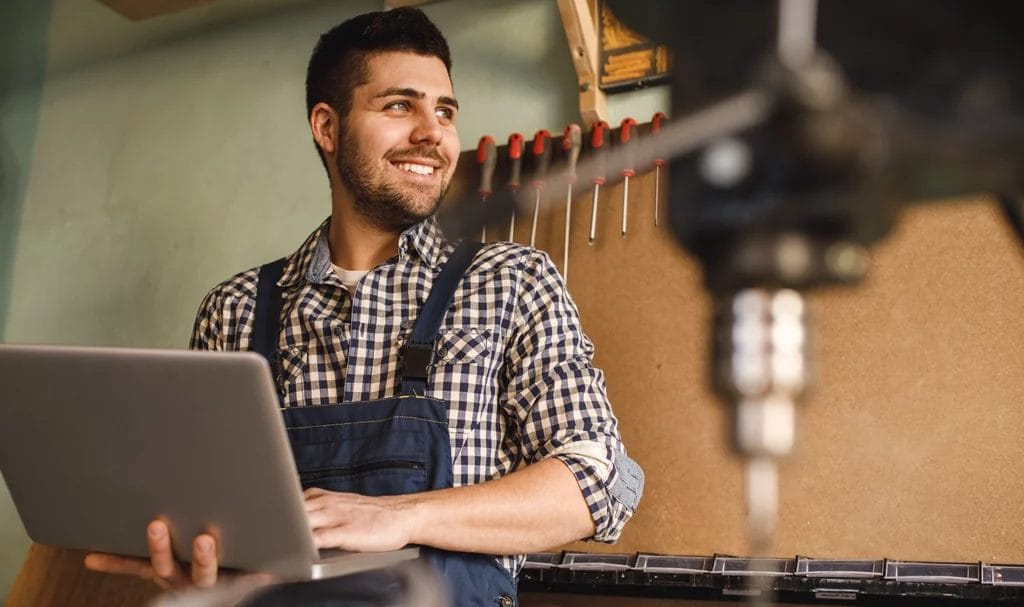 A craftsman smiles while using his laptop from his studio, where he makes and sells products.