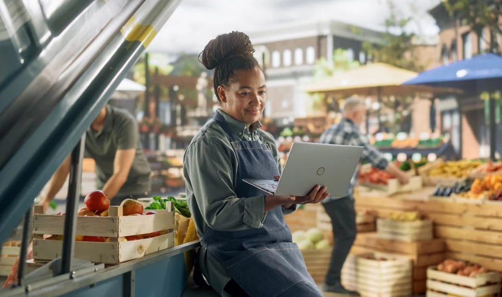 A farmers market vendor smiles at her laptop while she leans up against the side of her booth and works on business items before the market begins.