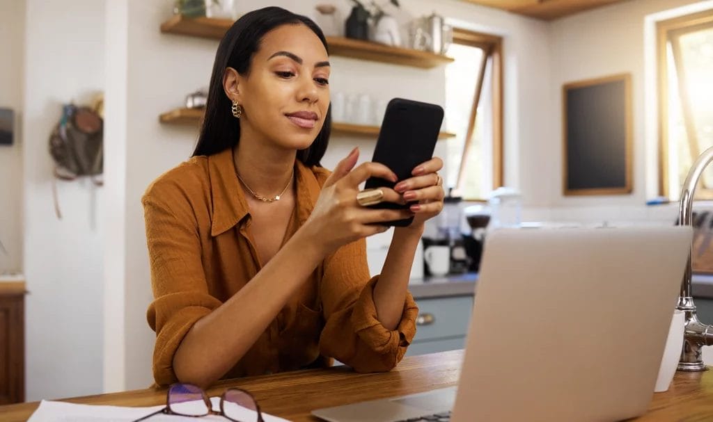 A small busines owner works from her home where she is using both her phone and laptop to complete business tasks online.