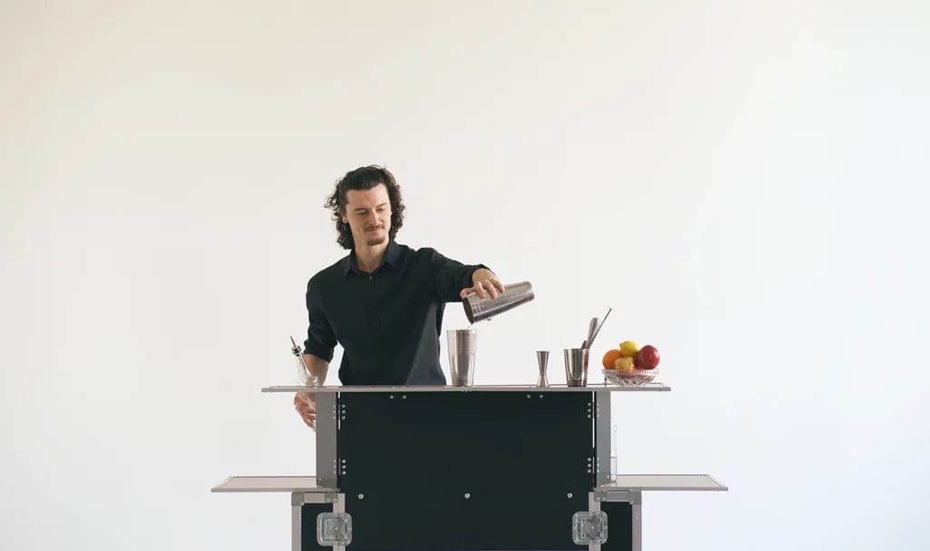 A professional bartender is preparing a cocktail on a mobile bar table against a white background.