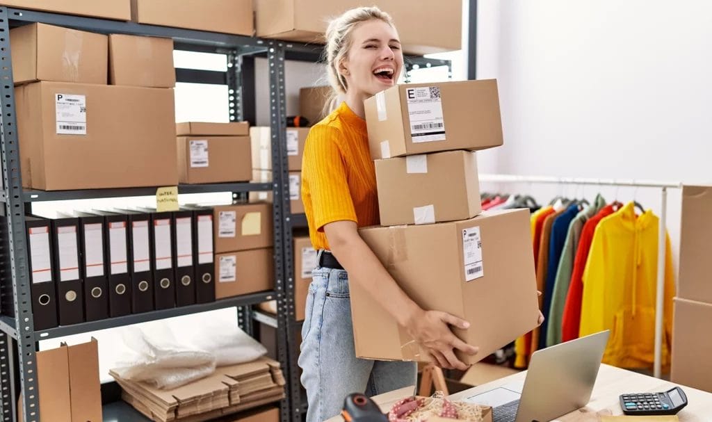 A young female business owner laughs as she carries a large stack of boxes from her workspace to be shipped to customers.