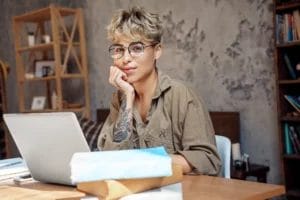 A woman with short wavy hair, wearing glasses and a taupe shirt, rests her chin on her hand and smiles while sitting at a desk with an open laptop, stacks of books, and papers.