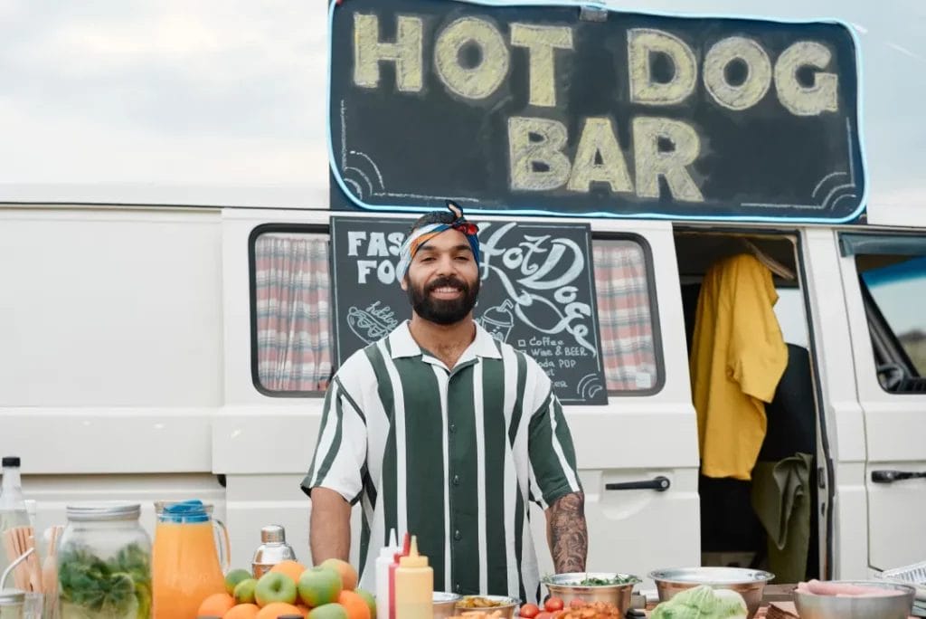 A man with a beard wearing a colorful bandana and a white and green stripe shirt smiles in front of a white food truck with a sign that reads, "Hot Dog Bar."