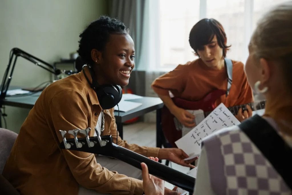 A woman wearing a brown shirt and headphones around her neck holds a paper with lyrics written on it and smiles at a fellow musician in a room.