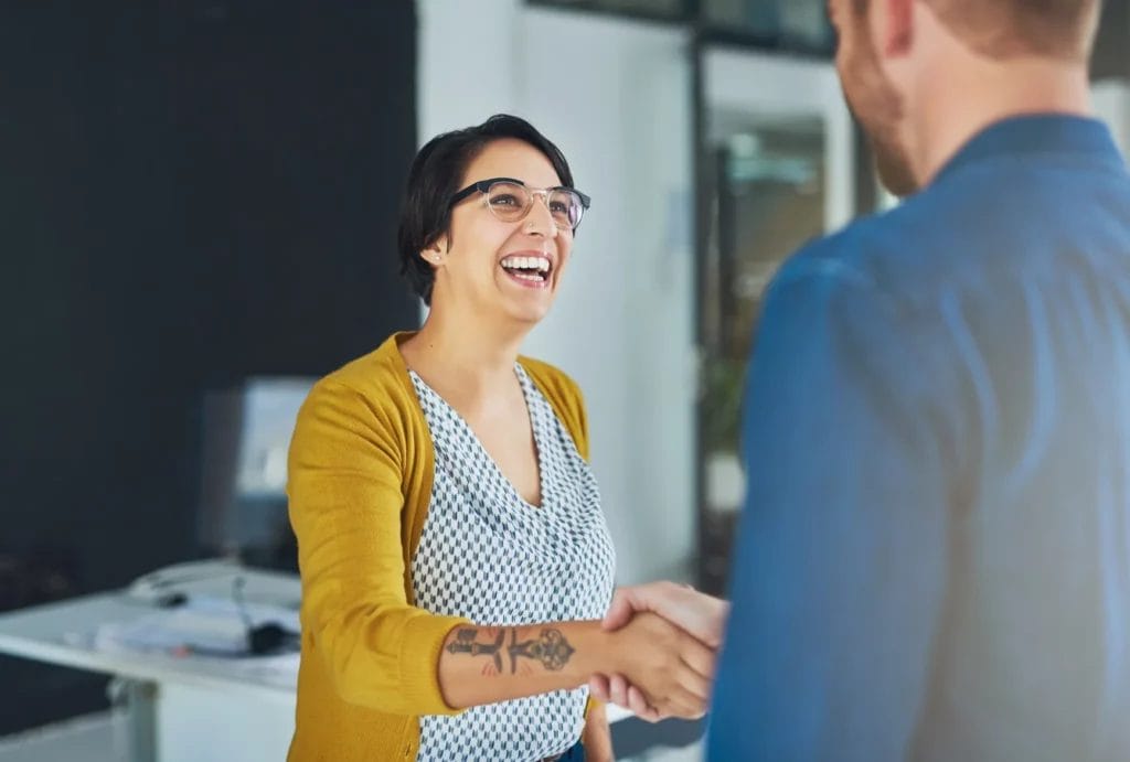 A woman wearing a white and blue printed top, a mustard cardigan, and glasses smiles while shaking hands with a man wearing a blue shirt in the foreground.