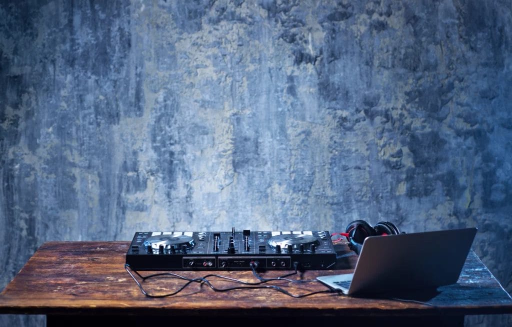A simple DJ set up featuring a black turntable, headphones, and an open gray laptop sitting on a wooden table in front of a blue-gray chalkboard background.