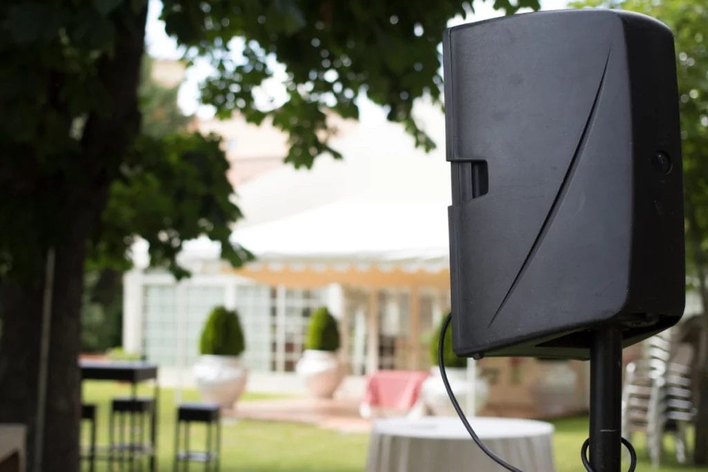 An asymmetrical shot of an outdoor DJ speaker setup with tables and chairs out of focus in the background.