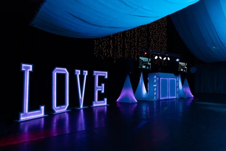 A blue and purple-lit wedding dance floor featuring large fluorescent letters spelling "love" and a DJ booth.