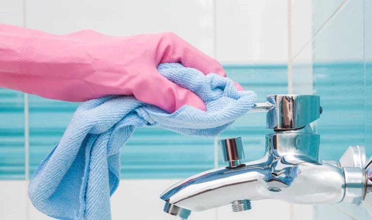 A close-up look of a cleaner with a pink rubber glove using a blue microfiber cloth to clean a bathroom sink handle.
