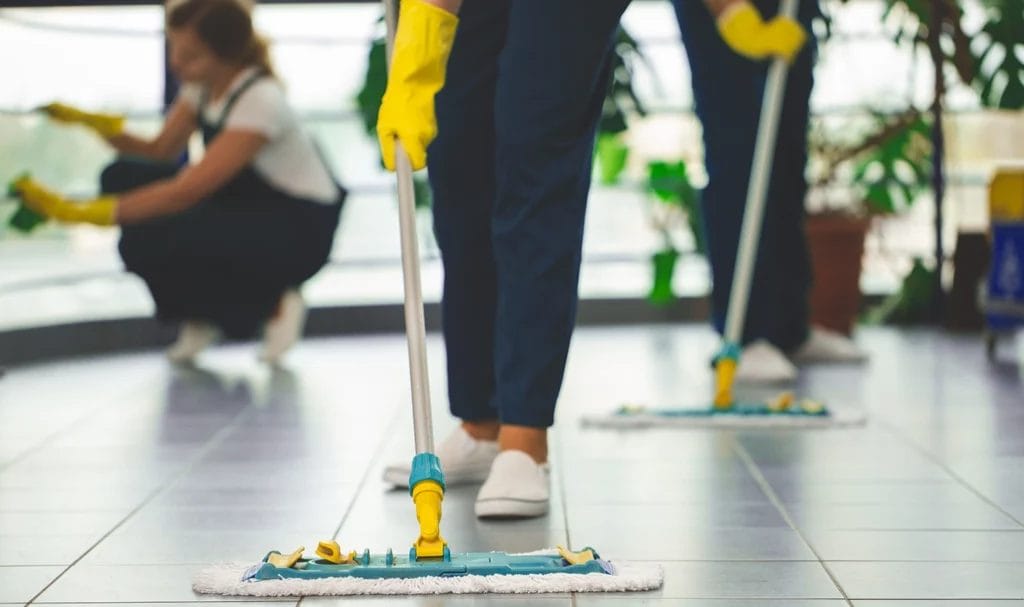 Close-up of a crew of professional cleaners with yellow gloves mopping and wiping the floors of a commercial office space.
