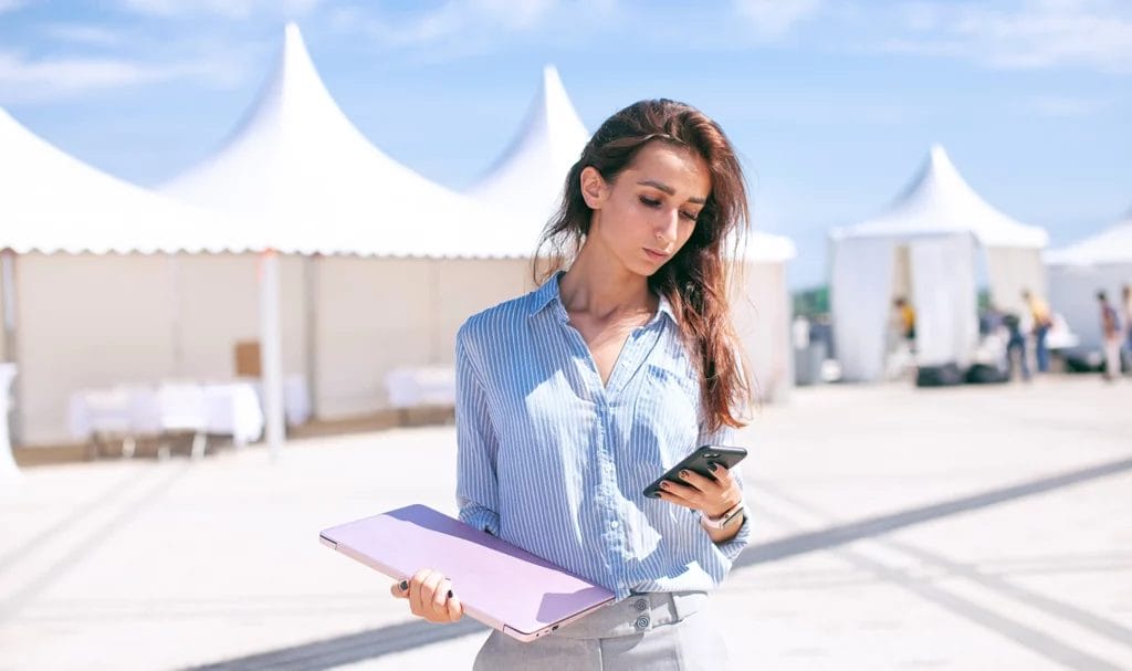 An event manager is preparing for an expo as she stands near empty vendor tents holding her smart phone and laptop.