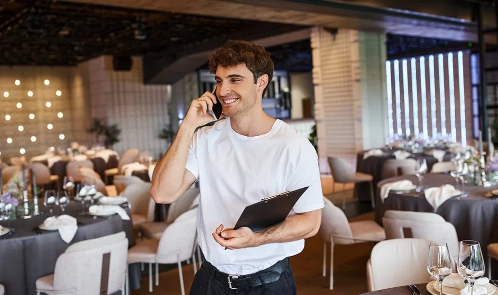 Smiling event organizer with clipboard talking on smartphone near festive tables in banquet hall.