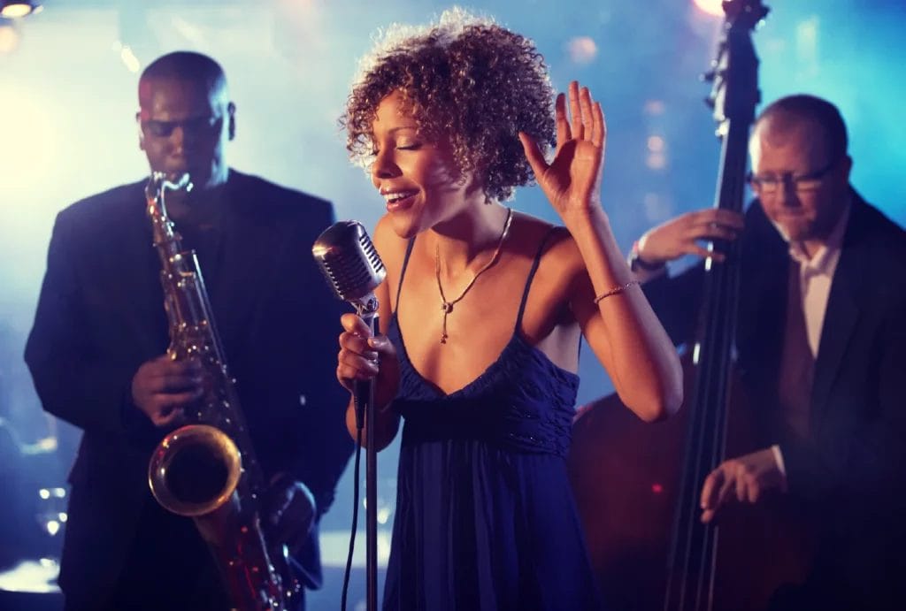 A jazz bands performs live, with the lead singer in focus in the middle and the saxophonist and bass player behind her.