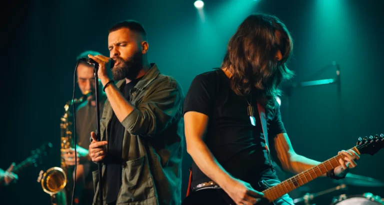 Rock band performing live on stage with a turquoise backlight, featuring a close-up of the lead singer in the middle, a saxophonist behind him, and the lead guitarist to the side.