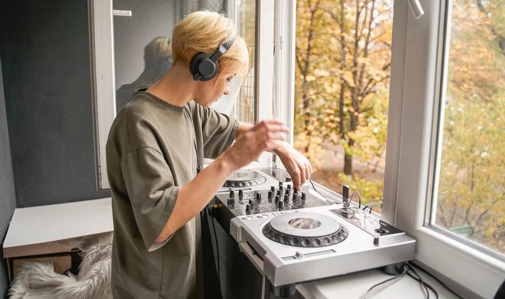 A young female DJ is enjoying mixing music in her home next to an open window.