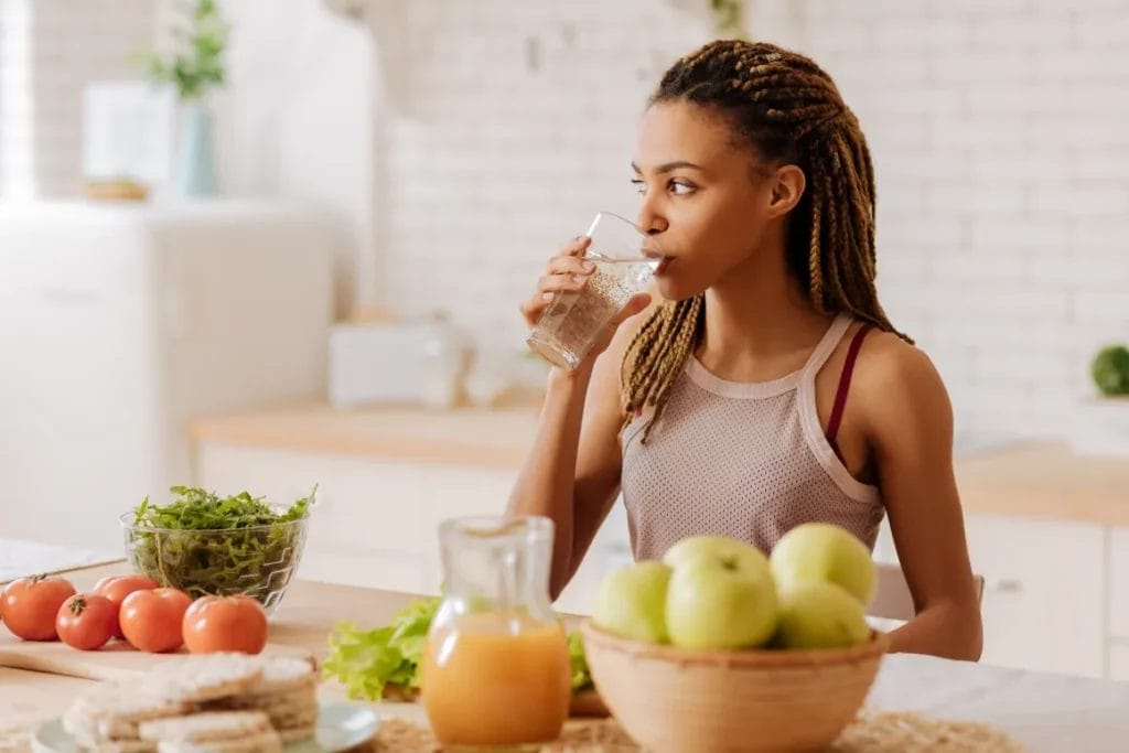 A woman drinks a glass of water in a white kitchen in front of a table with tomatoes, apples, a glass pitcher of juice, and other foods placed atop its surface.