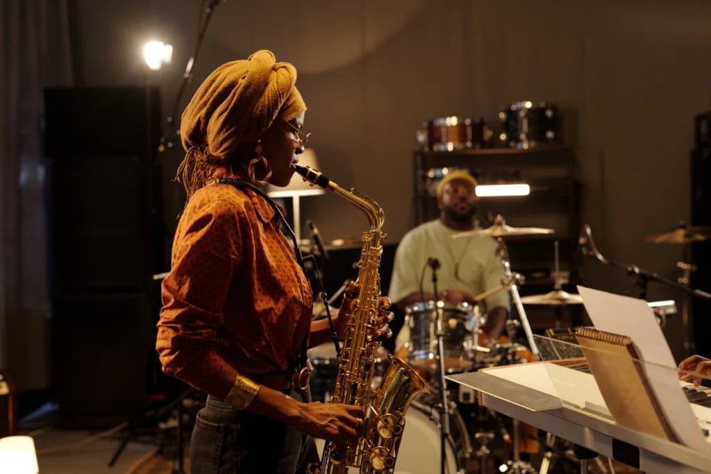 Musician in dark orange clothes plays the saxophone in the foreground, with a drummer in the background of a recording studio.