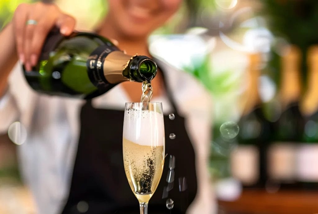 A woman in a white shirt and black apron pours champagne into a glass.