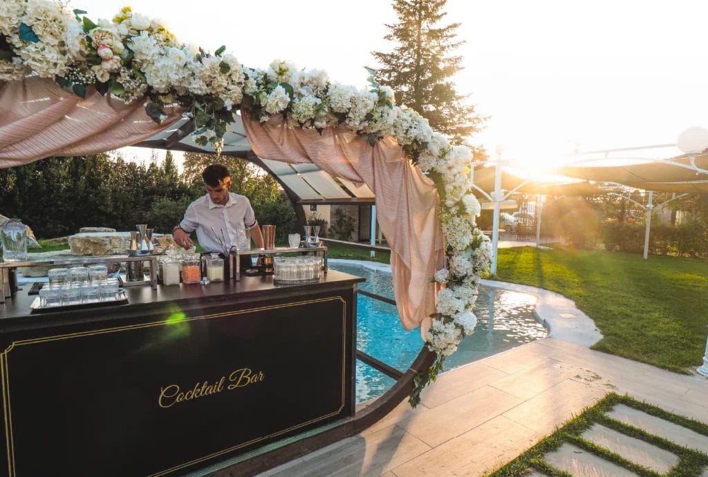 A young male bartender working at a cocktail bar next to a pool at an outdoor wedding.