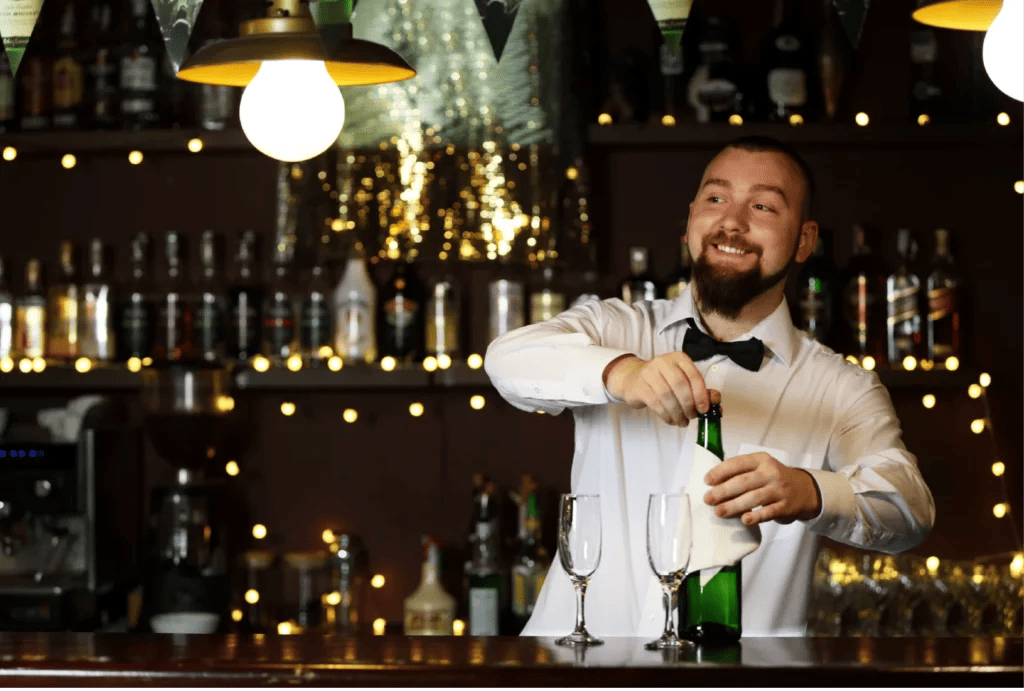 A male bartender in a white button-up shirt and black bowtie smiles while opening a bottle of champagne.