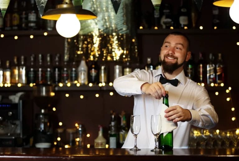 A male bartender in a white button-up shirt and black bowtie smiles while opening a bottle of champagne.