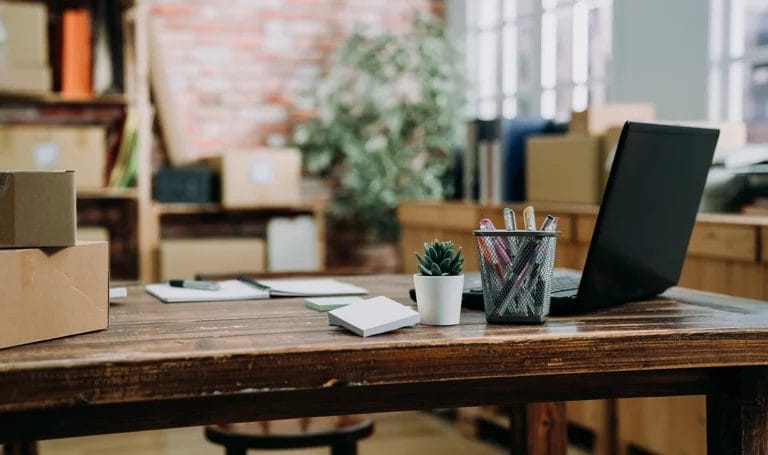 Open concept office space with a wood desk that has a laptop, pens, boxes, notebook, and other small business office supplies on it.