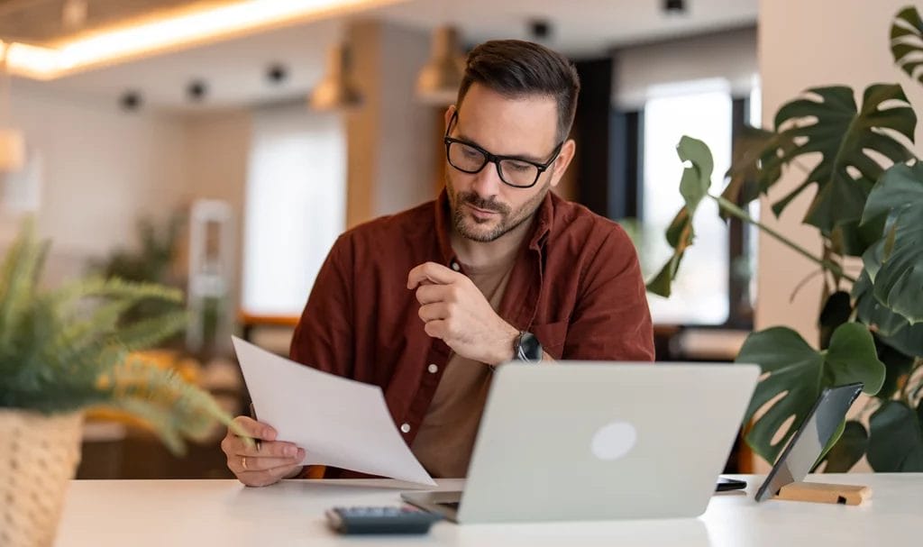 An entrepreneur using a laptop while sitting at a table in a home office and looking at paperwork.