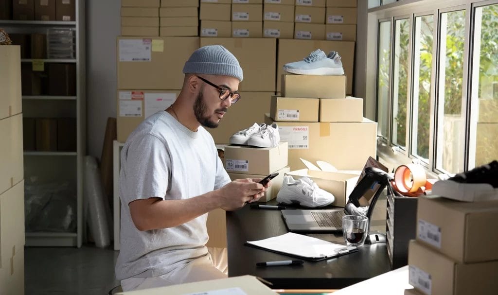 An online seller working from his phone and laptop in a small storage warehouse.