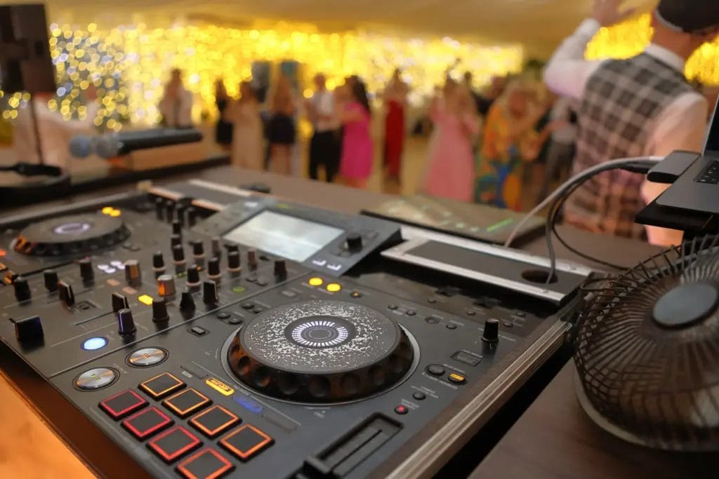 Close-up of a DJ table with out of focus wedding guests dancing in the background.