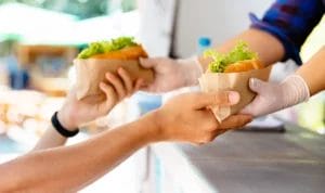 A close-up look of a food truck owner handing out burgers wrapped in wax paper to a customer.