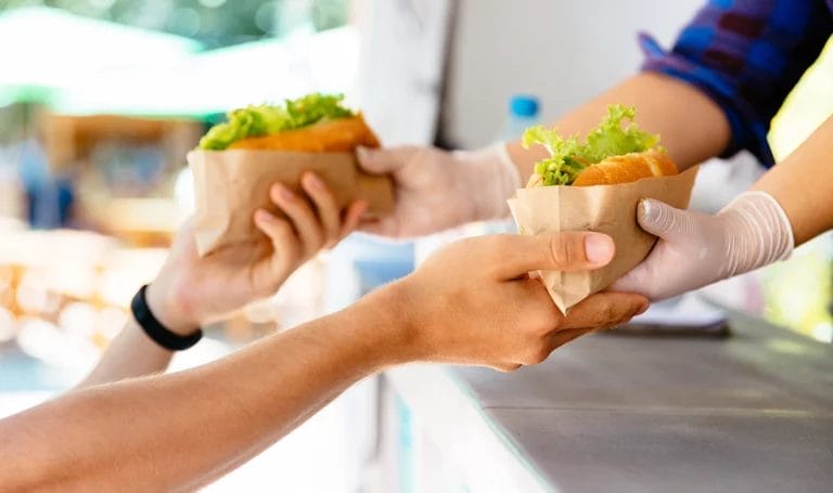 A close-up look of a food truck owner handing out burgers wrapped in wax paper to a customer.
