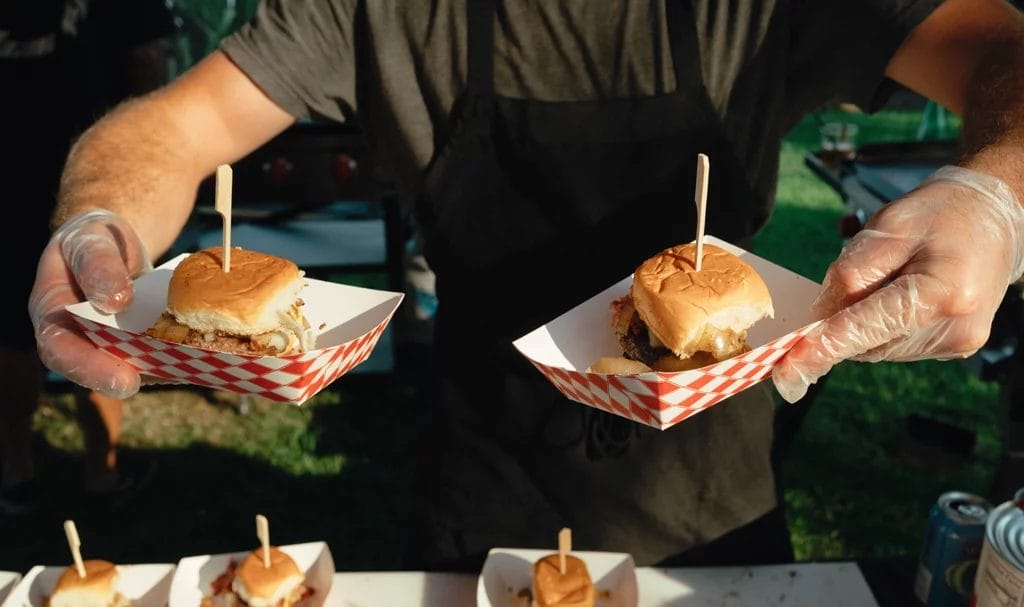 Close-up of a food vendor serving fresh sliders in checkered paper trays.