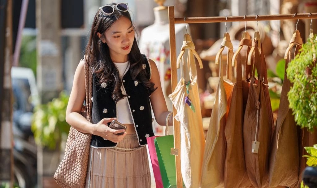 A shopper looks through handmade tote bags on display in a vendor booth at an outdoor market.