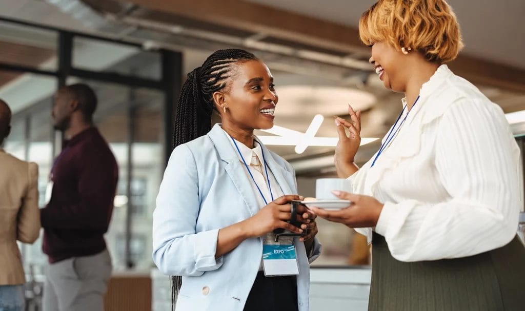 Professional women networking at a business expo, holding coffee and chatting.