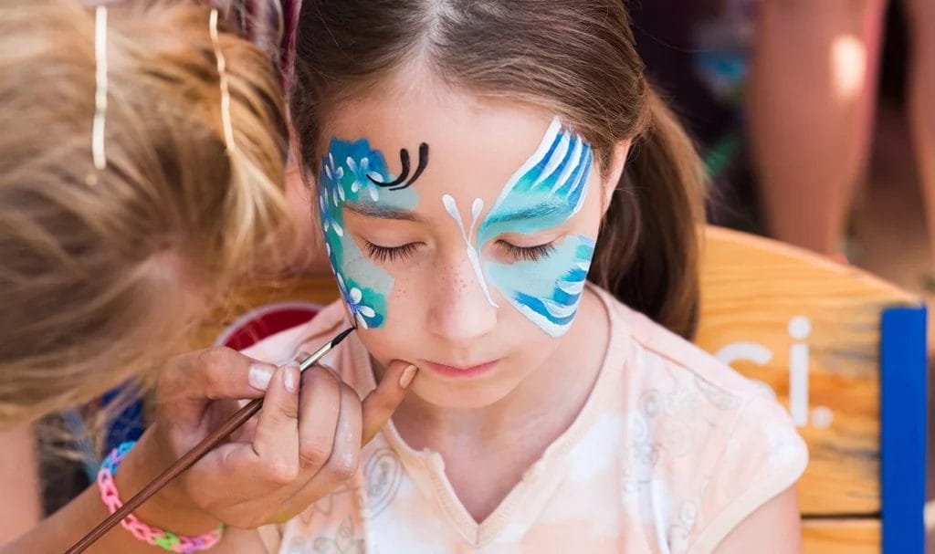 A face painter carefully brushes blue face paint on a young girl to create a butterfly.