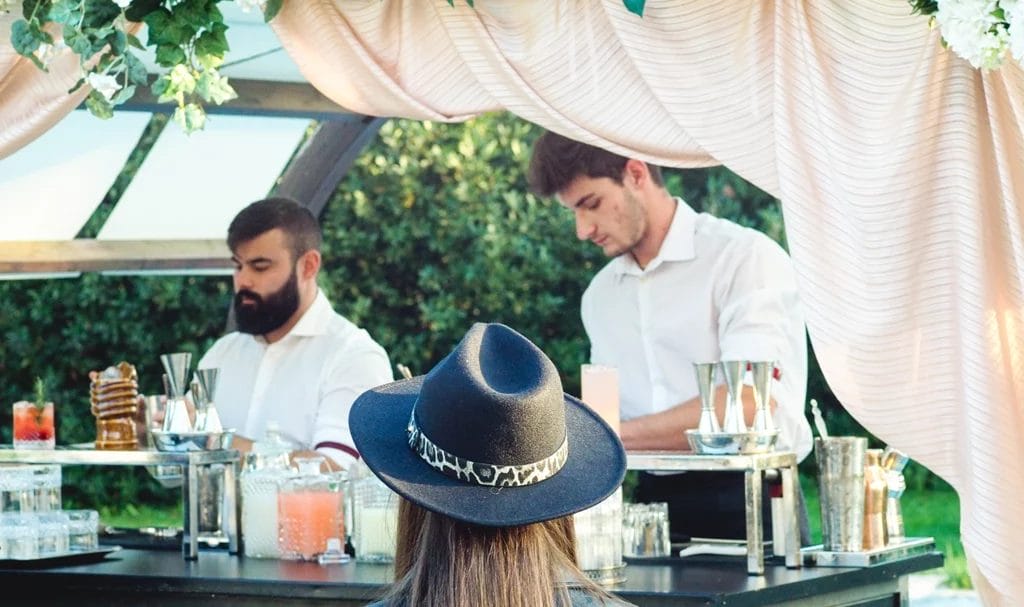 A mobile bar is set up at an outdoor wedding and is serving guests hand-mixed drinks.