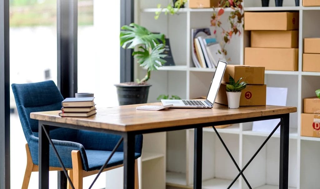 An online small business owner's home office with a laptop, ready to ship packages, a calculator, and a stack of books on a desktop.