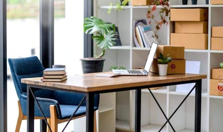 An online small business owner's home office with a laptop, ready to ship packages, a calculator, and a stack of books on a desktop.