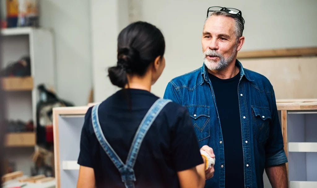 An older gentleman shakes hands with a young businesswoman in a small wood workshop.