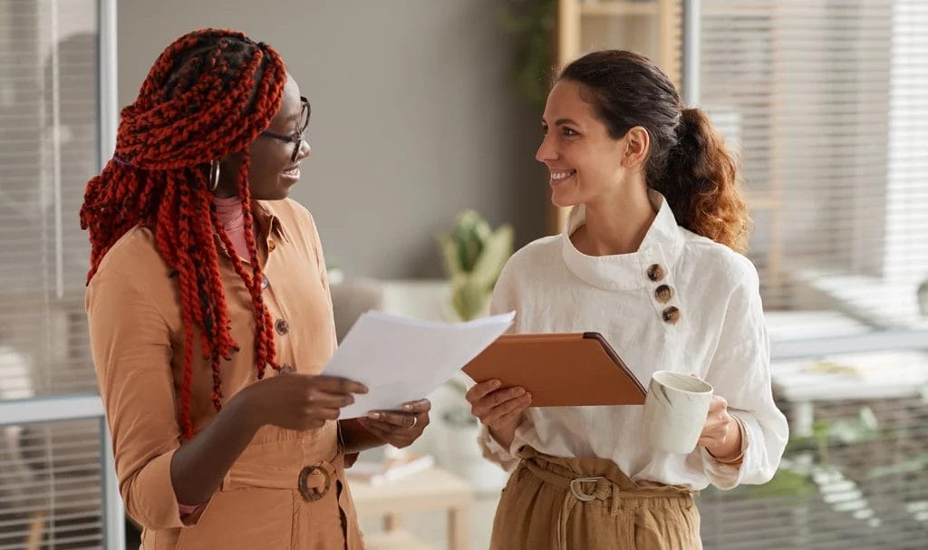 Two young businesswomen discussing insurance documents and smiling cheerfully while standing in modern office interior.