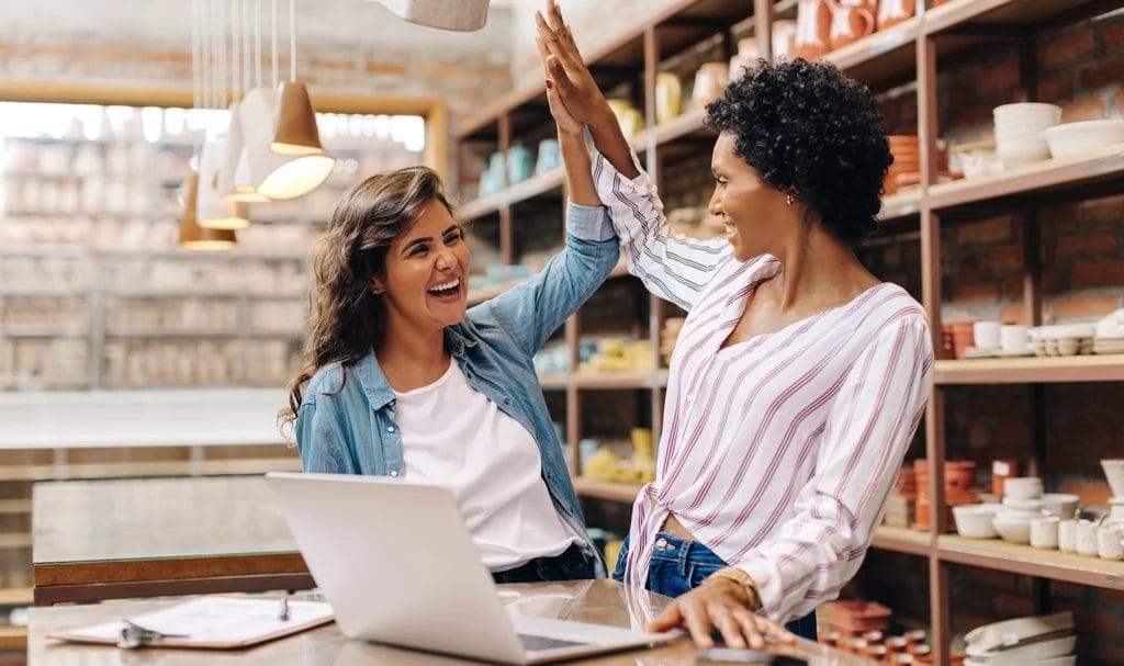 Two ceramic shop owners celebrate their success by high-fiving each other. A laptop is open in front of them, while a shelf full of handmade ceramic pieces is behdind them.