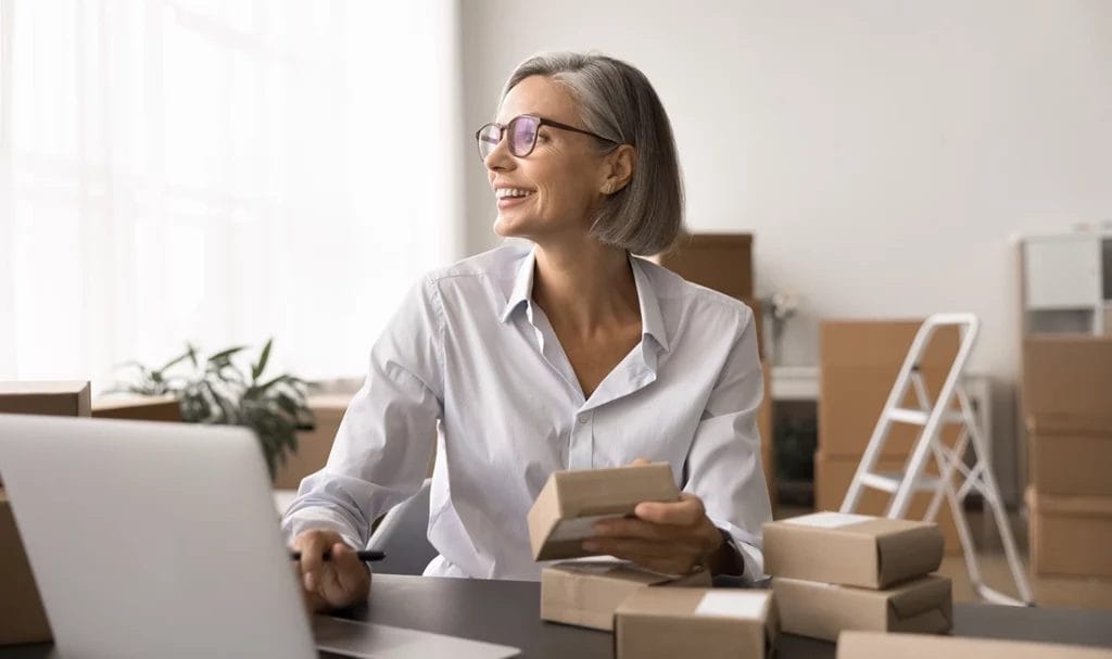 A happy, middle-aged small business owner is looking out the window with a contented smile as she sits at her desk with a laptop surrounded by cardboard boxes and various packages prepared for shipment in her workspace.